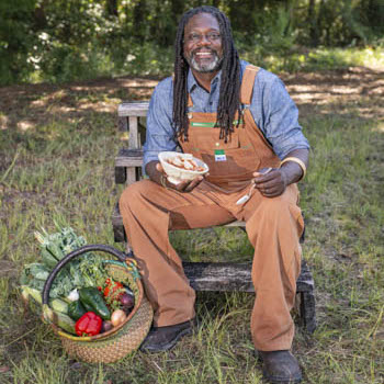 Matthew Raiford sitting in brown overalls eating a bowl of food with a basket fruits and vegetables by his feet.